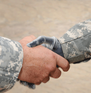 Foto de la portada: Una imagen centralizada de un apretón de manos entre dos miembros del servicio en uniformes del camuflaje.  La mano de una persona es una prótesis.