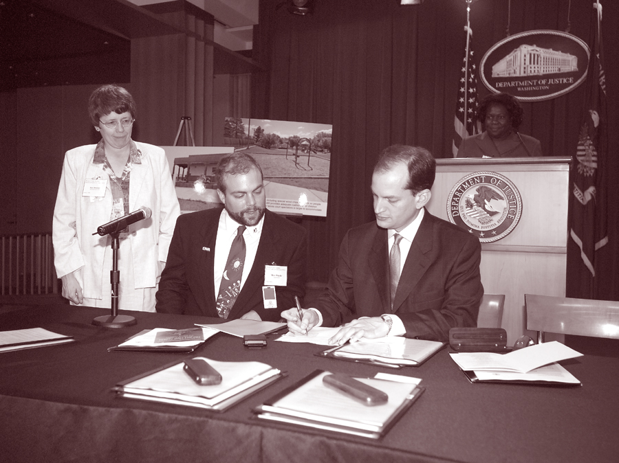 Photo of Former Assistant Attorney General R. Alexander Acosta signing PCA Agreement Between DOJ and Juneau, Alaska, at August 2004 DOJ ceremony with Deputy Assistant Attorney General Loretta King, and Building Inspector Sara Boesser and Assemblyman Marc Wheeler of Juneau, Alaska, looking on.