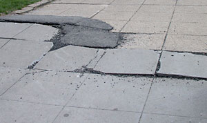 Photo of a sidewalk with broken pavement, raised sections, and patches