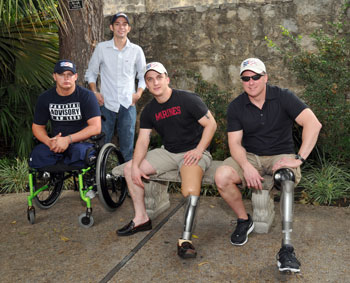 Photo:  Three seated men pose for the camera:  two of them sit on a bench and have prosthetic legs; one sits in a wheelchair and has double leg amputations.  A fourth man stands behind the group.  