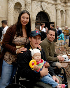 Photo: A man sits in a wheelchair with a woman standing behind him, her hands on his shoulders.  A baby sits in his lap.  
